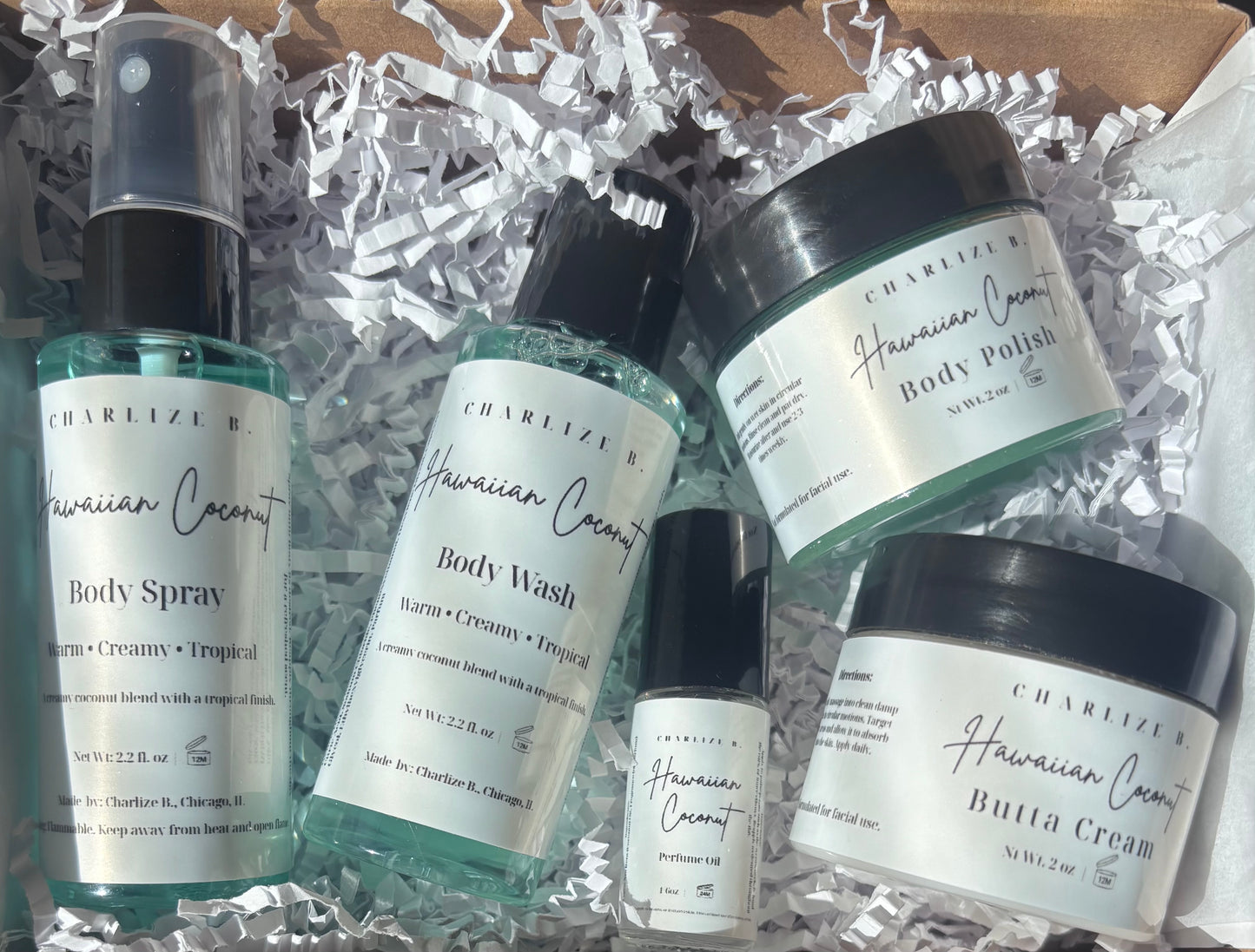 A set of five skincare products from Charelize K Hawaiian Coconut, including body spray, body wash, body polish, butta cream, and a small bottle, arranged in white crinkle paper inside a cardboard box.