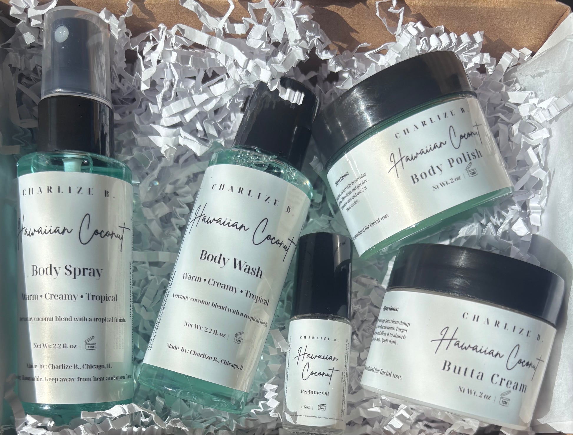 A set of five skincare products from Charelize K Hawaiian Coconut, including body spray, body wash, body polish, butta cream, and a small bottle, arranged in white crinkle paper inside a cardboard box.